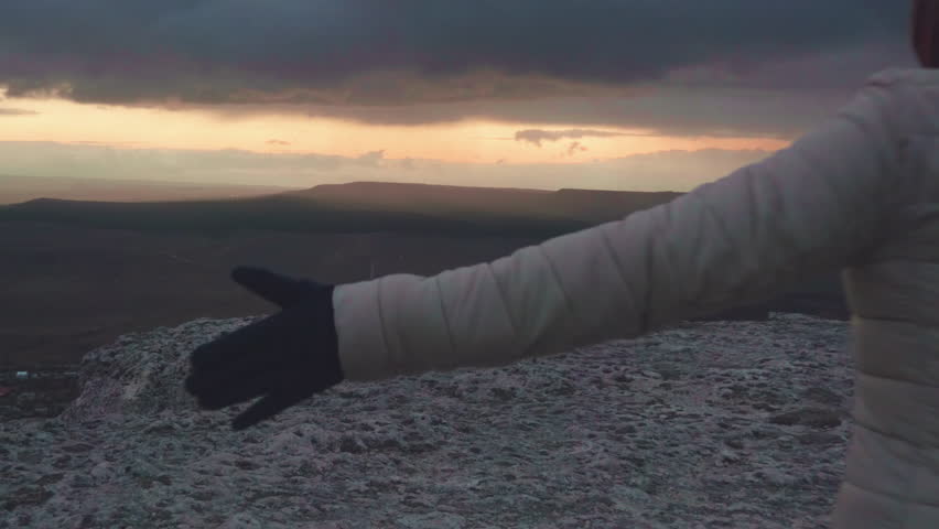 Young cheerful woman is running with raised her hands on the top of mountain. Wind flutters hairs. Dramatic sky at dusk. Slow motion.