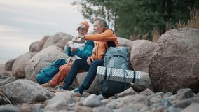 Senior man woman campers drinking hot drink from plastic cups talking enjoying landscape on riverbank. Old couple family pensioners travelling together. Travel tourism camping journey hiking concept. - Powered by Shutterstock - Get 15% off with code: PIKWIZARD15