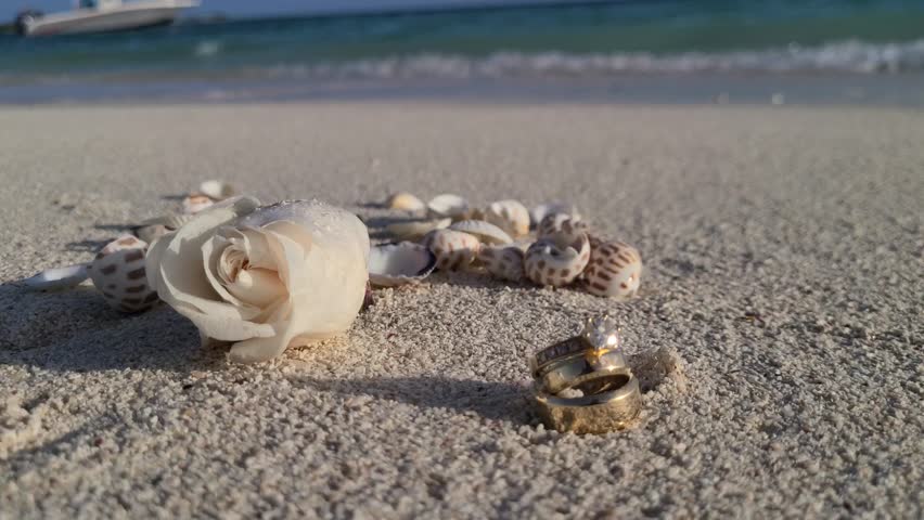 Single pristine white flower adorning a gleaming wedding ring on soft sand, sea background