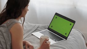 A long-haired brunette lies on a bed with a cup of tea and looks at something on her laptop. A woman lies in front of a laptop with a green screen and periodically presses the keys.