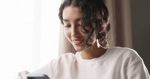 Bright sun pours into room, girl sits on sofa and scrolls through social networks. Lazy weekend, relax and enjoy peace and quiet. Girl smiles while chatting with her friends on Internet. Cinematic AD.
