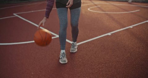Close-up a girl in a sports uniform hits an orange basketball ball from a red floor on a street court at sunrise in summer. The girl goes in for sports and develops the ability to play basketball inの動画素材