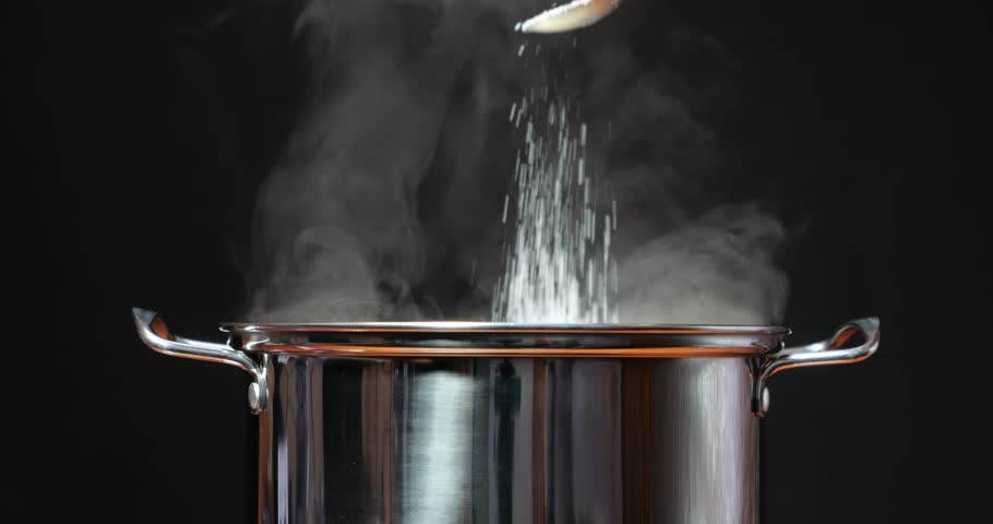 Steaming hot water in a steel pan. Salt the water with your hand or a spoon. Cook dinner and food in the kitchen by adding seasoning. Steam rises from boiling water against a black background.