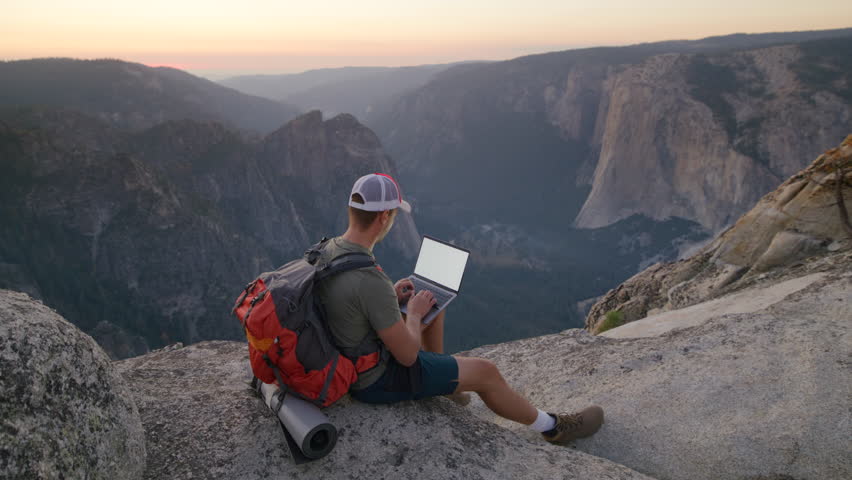 A hiker sits on a mountain edge with a laptop, taking in the amazing view of Yosemite Valley as the sunset paints the sky. Slow Motion, Camera 4K RAW.  Royalty-Free Stock Footage #3430424715