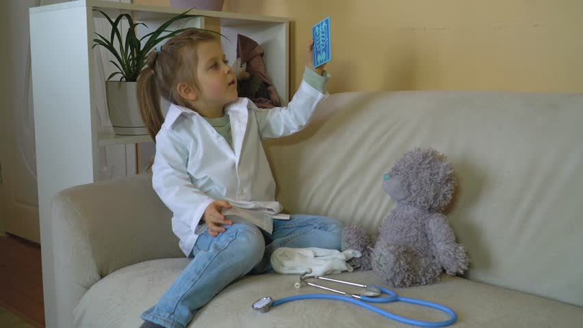 Caucasian girl in white medical coat with stethoscope treats teddy bear, looks X-ray, playing hospital, playing nurse, veterinarian Happy family, role-playing games, children's professions Royalty-Free Stock Footage #3430693921