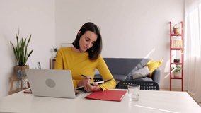 Young adult student woman taking notes while using laptop computer at home. Millennial pretty female learning online listening virtual video call. Business and education concept.
