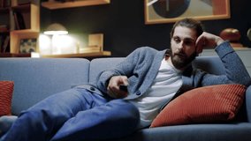 Portrait of tired young bearded man with remote controller switches channels on TV trying to choose something interesting looking at screen at late night. Boring TV shows films movie