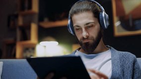 Young handsome man in headphones watching video stream on modern tablet while sitting on sofa in living room at home. Smiling male watches online broadcast, or plays computer video game. - Powered by Shutterstock - Get 15% off with code: PIKWIZARD15