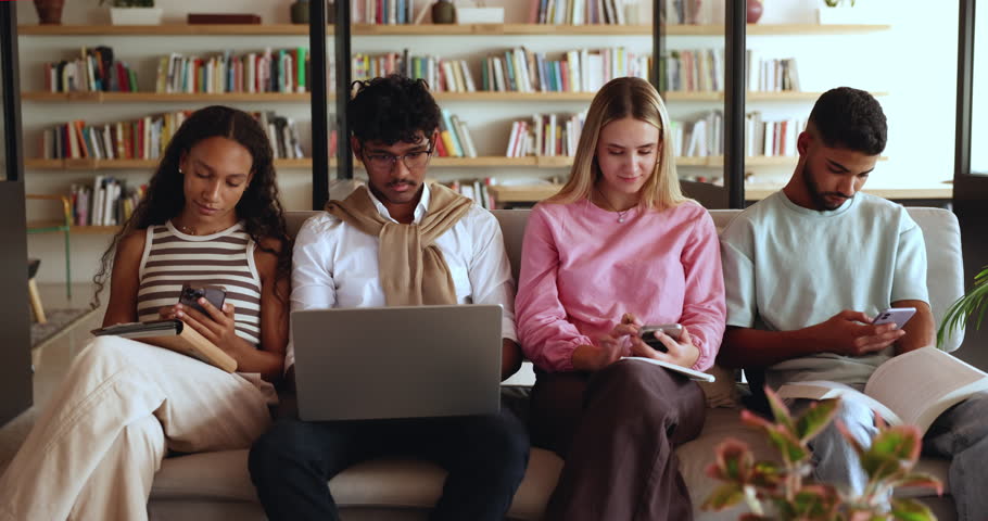 Gen Z students using diverse modern wireless devices in library, distracted from study, chatting, check schedule via mobile on-line apps looks focused ignoring each other, absorbed in internet usage