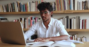 Serious young Indian male student studying with computer in library, communicate with friend by videocall app on laptop, discuss new university tasks, prepare for exam. Distance learning, education