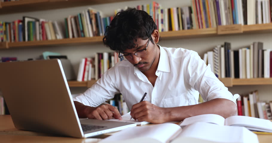 Focused Indian student guy sit at desk in library writing essay, making notes in workbook, learning theory, gaining new knowledge, e-learning using computer application. Modern tech, education, skills Royalty-Free Stock Footage #3430811605