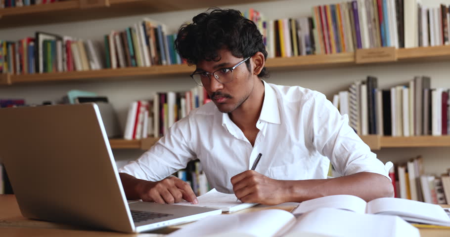 Focused Indian student guy sit at desk in library writing essay, making notes in workbook, learning theory, gaining new knowledge, e-learning using computer application. Modern tech, education, skills Royalty-Free Stock Footage #3430811605