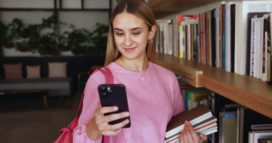 Pretty young student girl standing in university library, holding mobile phone, communicate with classmates, send message, check class school schedule, take break, web surfing. Modern tech, education Royalty-Free Stock Footage #3430813341