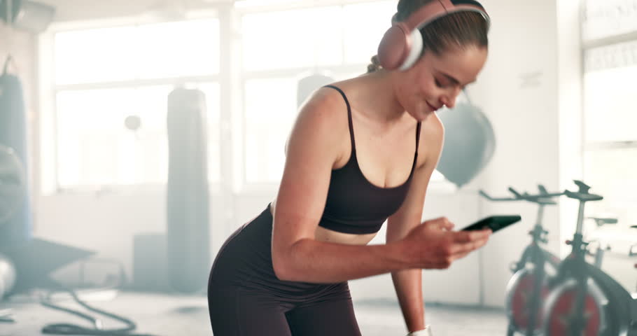 Happy woman, headphones and phone on break at gym for music, podcast or social media. Fitness, young female person or athlete in rest, relax or listening to sound on mobile smartphone at health club