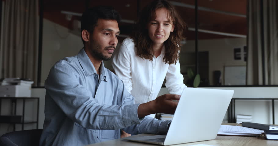 Positive diverse business colleagues discussing online project at laptop, looking at display, talking at workplace. Young Indian mentor man training Caucasian intern girl, pointing at screen, speaking Royalty-Free Stock Footage #3430877629