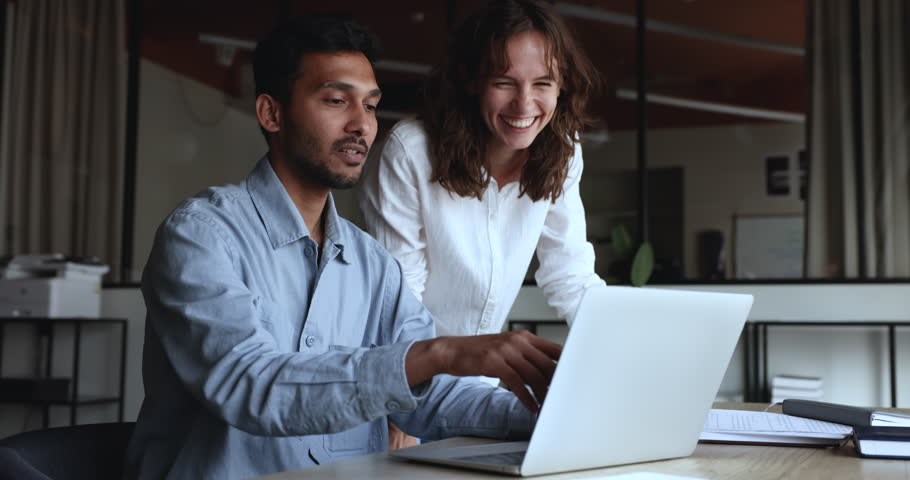 Positive diverse business colleagues discussing online project at laptop, looking at display, talking at workplace. Young Indian mentor man training Caucasian intern girl, pointing at screen, speaking