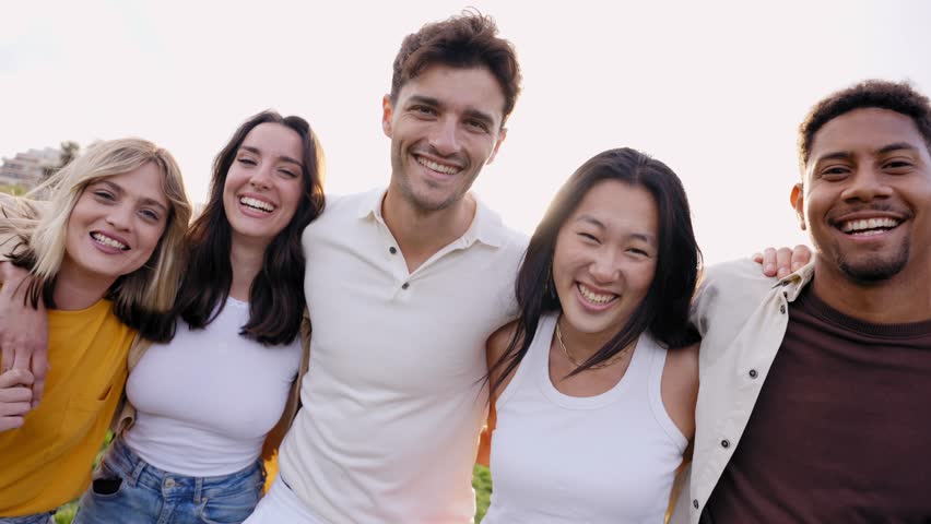 Multirracial group of young smiling friends looking at camera in the park hugging in community. Happy millennial people laughing taking selfie having fun together outside. Cheerful university students