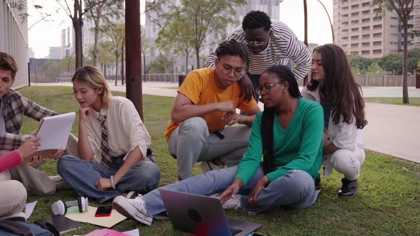 Group of young international university students sitting on the grass outside the faculty building. Friends gathered happily studying together with workbooks and laptop on campus