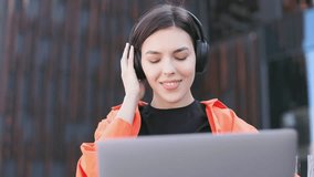 Portrait of smiling woman enjoying music in headphones and nodding head to rhythm while sitting with laptop outdoor. Aspiring female musician watching personal video clip on streaming platform.