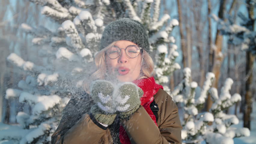 Beautiful young woman with glasses blowing snow from palms and smiling to camera, portrait in park. Pretty girl resting in forest in winter day, good mood and positive emotions, happy Christmas day