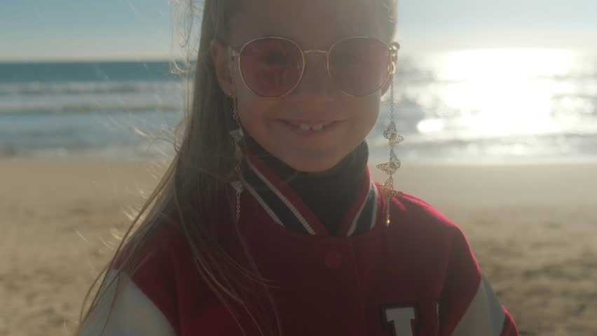 Portrait of a little happy smiling girl wearing sunglasses. Spring sunny day on the beach against the backdrop of the sea.