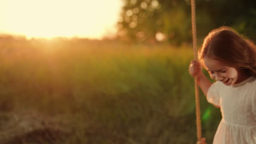Child plays on wooden swing, dreams of fly. Child swing, kid girl smile in flight. Child girl Happiness dream entertainment. Happy little girl swings on swing in park under tree at sunset. Family walk