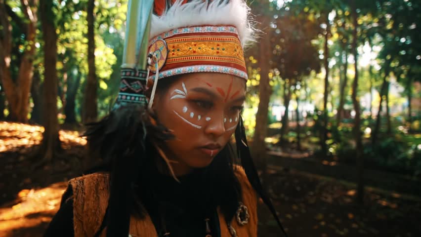 Portrait of a person in traditional indigenous attire with face paint, standing in a forest setting in the morning