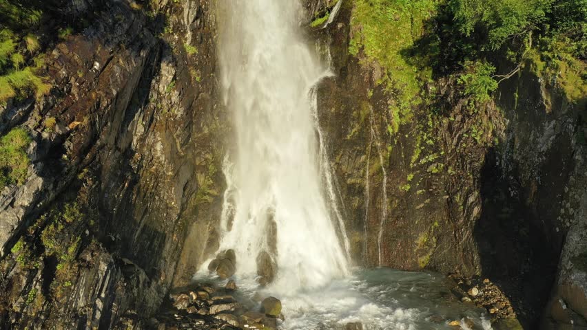 Large streams of waterfall among the branches of trees fall down in close-up. A smooth ascent above the stream among the trees at sunset.