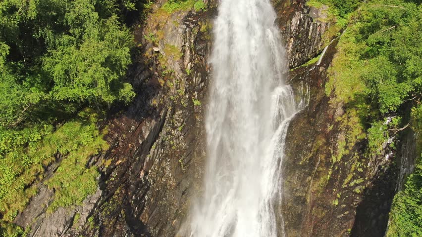 A smooth ascent over the stream of a large waterfall among branches and trees at sunset on a sunny day.