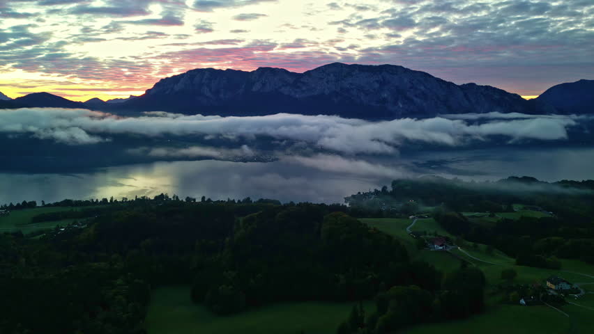 Colorful sunrise beyond the silhouettes of Alps in Austria with morning clouds over a lake. Aerial view