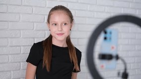 Beautiful teenage girl posing in front of a phone on a tripod. Child blogger shoots video in front of camera and ring lamp
