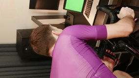 Male is training on exercise bicycle. Man is cycling on stationary smart trainer and looking at green screen. Cyclist is pedaling on bicycle simulator. Vertical video