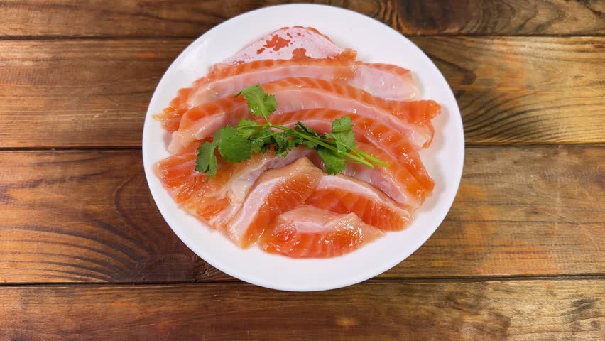 Salted salmon bellies strips without skin and fresh cilantro twigs on the white dish on a rustic table, view close-up while rotation
