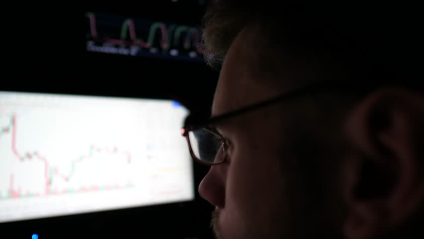 Young man works on computer at night. Profile of broker analysing real-time stocks commodities and foreign exchange charts. Financial analyst trading online at stock market. Concept of freelance job