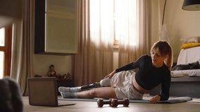 Resilient woman practices side plank with her prosthetic leg on an exercise mat. She follows a streaming fitness class, surrounded by dumbbells on the floor. Showcases strength, adaptability, and fitn