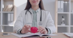 Close up portrait of a female smiling woman doctor cardiologist sitting at the desk on her workplace holding red heart in medical clinic. Health care, cardiology concept. 4K video.