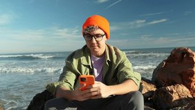 Attractive smiling young man happy using phone sitting on a rock on the beach. Communication travel man chatting scrolling social media. Close up. Slow motion. - Powered by Shutterstock - Get 15% off with code: PIKWIZARD15