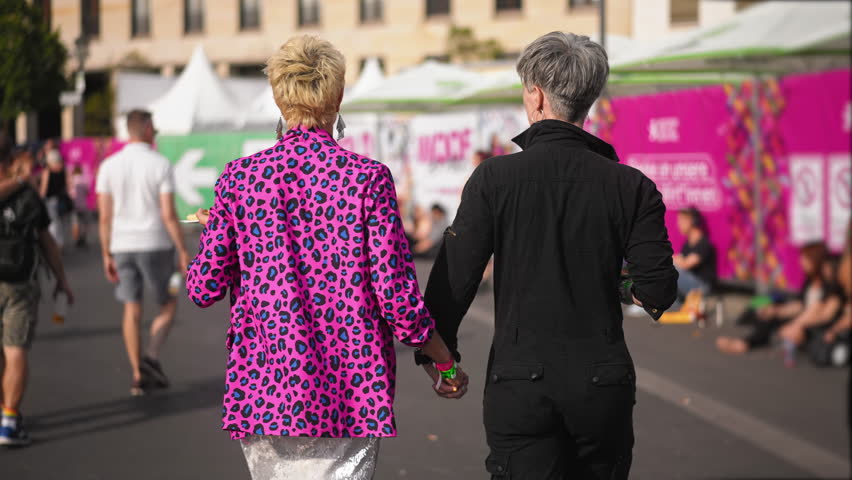 Happy lesbian couple hold hand walk city street. Feminism csd day. Dressed up middle aged lady. Same sex lover coming out. Bi feminist women rest have fun. Two gay girls love. Free feminism lgbt pride