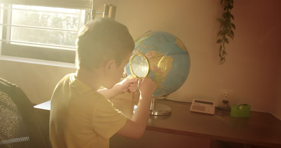 Child is studying while sitting at home in rays sunset in room. Kid in morning light sunset, cute little boy looking at globe through a magnifying glass, technology for schoolchildren. 4K