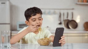 Indian preteen kid eating noodles while using mobile phone at home on dining table - concept of unhealthy lifestyle, internet addiction and technology