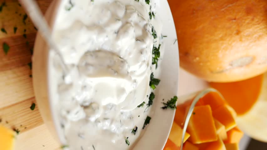 Vertical video: Adding fresh chopped herbs to heavy cream while making creamy pumpkin sauce. Close-up of a white bowl filled with sour cream and pieces of greens. Woman's fingers drop greens