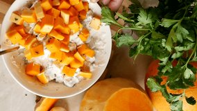 Top view woman mixes orange pieces of ripe pumpkin in a white thick creamy garlic sauce with herbs in a bowl using a spoon. Vegetable food made from fresh vegetables. Festive dinner