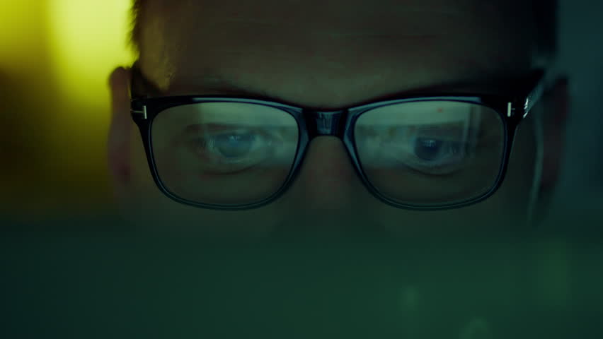 A laptop computer illuminates the face of a bespectacled programmer, who is intently analyzing the information on the screen. Information lines from the computer monitor are reflected in his eyes Royalty-Free Stock Footage #3432296647