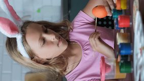 Vertical video. Child girl in bunny ears painting Easter eggs at table in the kitchen, slow motion