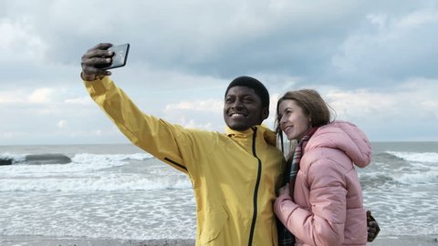 Two multicultural persons of black guy and caucasian female make a photography at smart phone close up. Positive fun concept of relations at windy spring vacation, cellphone take a pic with seaside