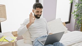 Handsome young man with beard laughing during a video call on a laptop in a bright bedroom.