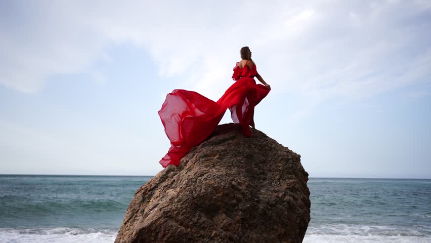 Red dress sea woman. Happy woman with flowing hair in a long flowing red dress stands on a rock near the sea. Travel concept, photo session at sea Royalty-Free Stock Footage #3432401193