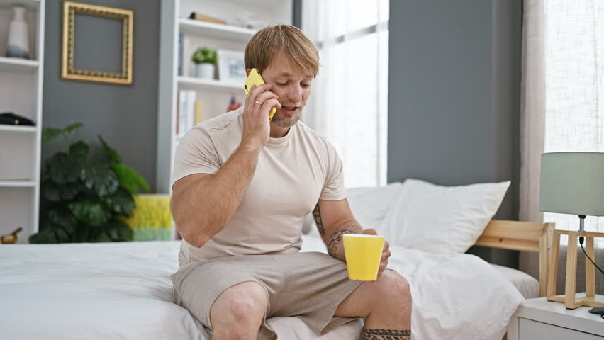 A bearded young man in casual clothes talks on a yellow smartphone while holding a mug, sitting on a bed in a well-decorated, contemporary bedroom.
