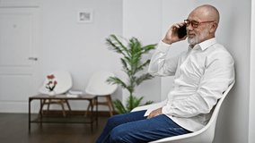 A senior man in a white shirt and glasses talks on the phone while sitting in a modern waiting room with white chairs and a decorative plant. - Powered by Shutterstock - Get 15% off with code: PIKWIZARD15