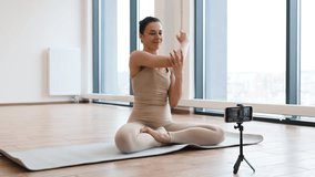 Sporty adult doing padmasana exercise on mat shooting video content on smartphone during yoga class. Side view of senior lady blogger in sportswear meditating in cross-legged sitting pose in studio.